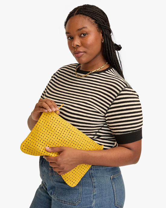 A woman in a striped t-shirt and jeans holds a Clare V Flat Clutch w/ Tabs in Dandelion Rattan, looking at the camera with a mild expression.