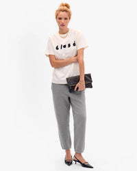 A woman stands against a plain background wearing a white T-shirt with classé, gray sweatpants, black pointed shoes, gold jewelry, and holds the Clare V. Foldover Clutch w/ Tabs - Petit Caviar in Black crafted from Italian leather.