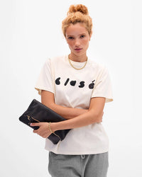 A woman with curly hair in a bun wears a cream clásé t-shirt, gray pants, gold necklace and bracelet, and holds the Clare V. Foldover Clutch w/ Tabs - Petit Caviar in Black, standing against a plain white background.