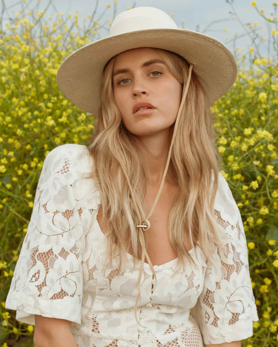 Woman wearing a white lace outfit and a Freya Field Gardenia in Bone hat standing in front of yellow flowers.