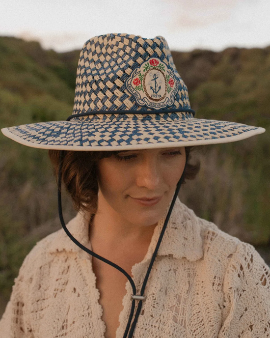 Woman with a subtle smile, wearing a textured white sweater and a wide-brimmed Sally Sells Lifeguard in Navy hat made of sustainable paper-based straw, outdoors with natural background. (Brand: Freya)