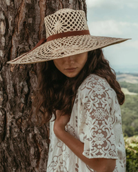 A woman wearing a wide-brimmed Freya Panama hat in Meringue/Natural/Brown and a lace-patterned top leaning against a tree.