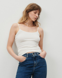 Woman in a American Vintage Gamipy Tank in Blanc and blue jeans standing against a plain background.