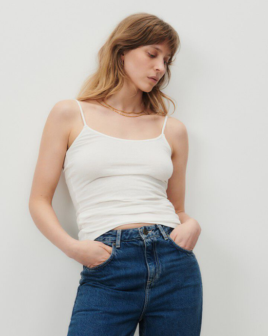 Woman in a American Vintage Gamipy Tank in Blanc and blue jeans standing against a plain background.
