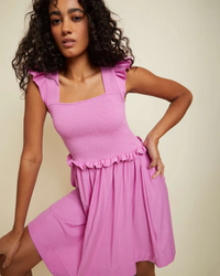 A woman posing in a pink sleeveless Nation LTD Ginger Smocked Mini in Prom Date dress with a square neckline and a smocked bodice.