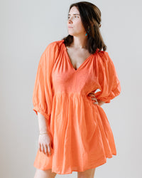 A woman in an orange Erin Shirred Waist L/S Dress in Dolly by Velvet by Graham & Spencer, posing with her head turned to the side.