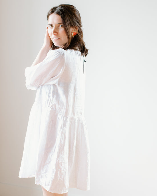 Woman in Velvet by Graham & Spencer's Kailani 3/4 Puff Slv Dress in White standing against a light background, looking over her shoulder with a gentle smile.