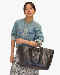 Woman in a denim jacket and leopard print skirt holding a large Clare V. Grande Bateau Tote in Black New Look, accessorized with layered necklaces.