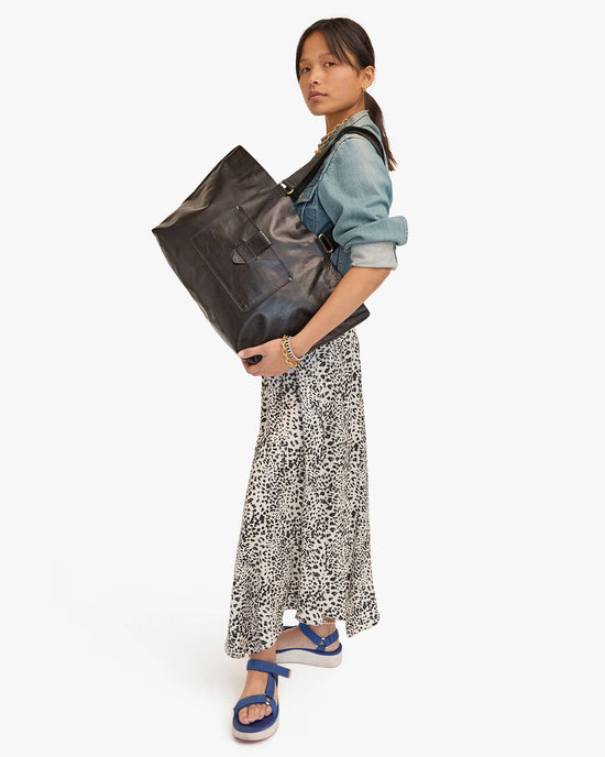 A woman stands in profile, wearing a denim jacket, printed skirt, and blue sandals, carrying a large Clare V. Grande Bateau Tote in Black New Look.