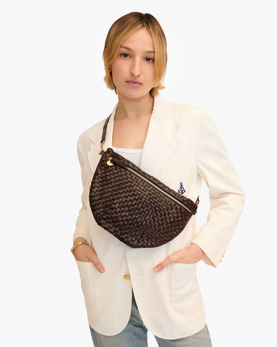 A woman in a white blazer holding a Clare V. Grande Fanny in Kalamata Woven Checker shoulder bag against a plain background.