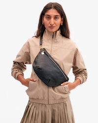 A woman in a beige jacket and pleated skirt stands with her hands in her pockets, modeling the Clare V. Grande Fanny - Petit Caviar in Black against a plain white background.