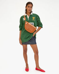 A woman in a green polo shirt and denim shorts, accessorized with a Clare V. Grande Fanny in Cuoio Perf bag, stands against a plain white background.