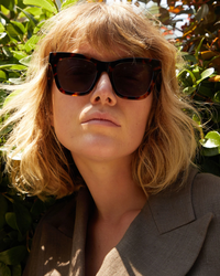 A person with blonde hair wearing Clare V. Heather Sunglasses in Tortoise with 100% UVA/UVB protection and a brown blazer, standing in front of a leafy green background.