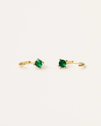 A pair of JaxKelly Emerald Huggies with green gemstones on a white background.