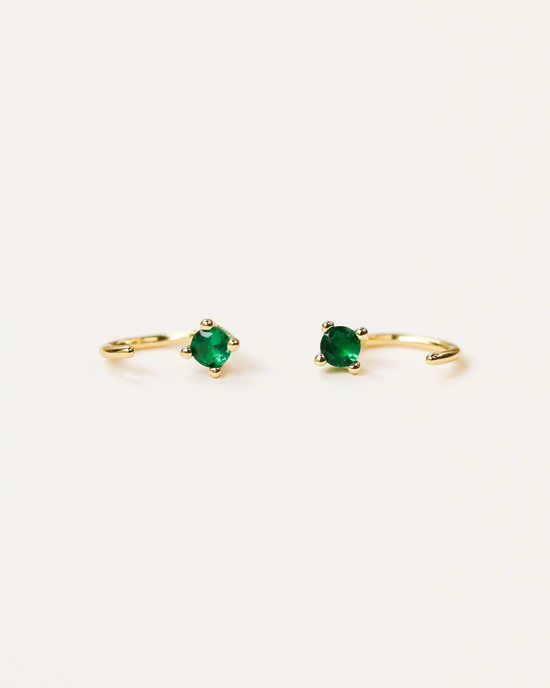 A pair of JaxKelly Emerald Huggies with green gemstones on a white background.
