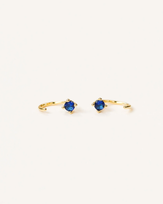 A pair of Sapphire Huggies with blue CZ stones on a white background by JaxKelly.
