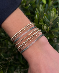 A person wearing multiple Karen Lazar Design 2MM Sig Bracelets with Envy Ombre & Yellow Gold beads on their wrist, against a backdrop of green foliage.