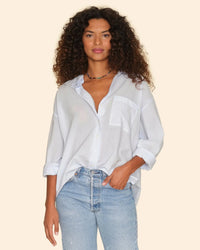 A woman with curly hair wears the XiRENA Sydney Shirt in Skylight with rolled sleeves and light blue jeans. She stands against a beige background, looking at the camera with a relaxed expression.