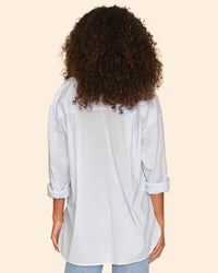 A person with curly hair, seen from behind, wears the XiRENA Sydney Shirt in Skylight with rolled-up sleeves and light blue jeans, standing against a beige background.