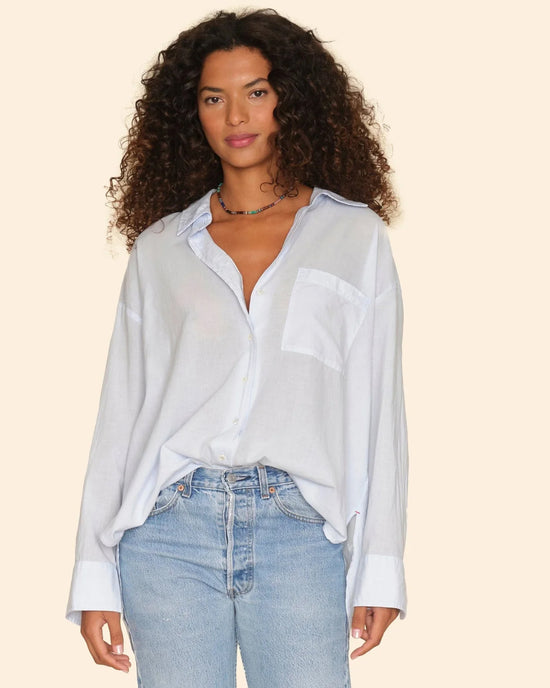 A woman with long curly hair models the XiRENA Sydney Shirt in Skylight, an oversized light blue cotton poplin button-down, partially tucked into jeans against a plain beige background.