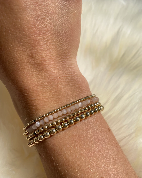 A wrist adorned with multiple 2MM Sig Bracelet with Nude Moonstone & Yellow Gold bracelets by Karen Lazar Design against a furry background.