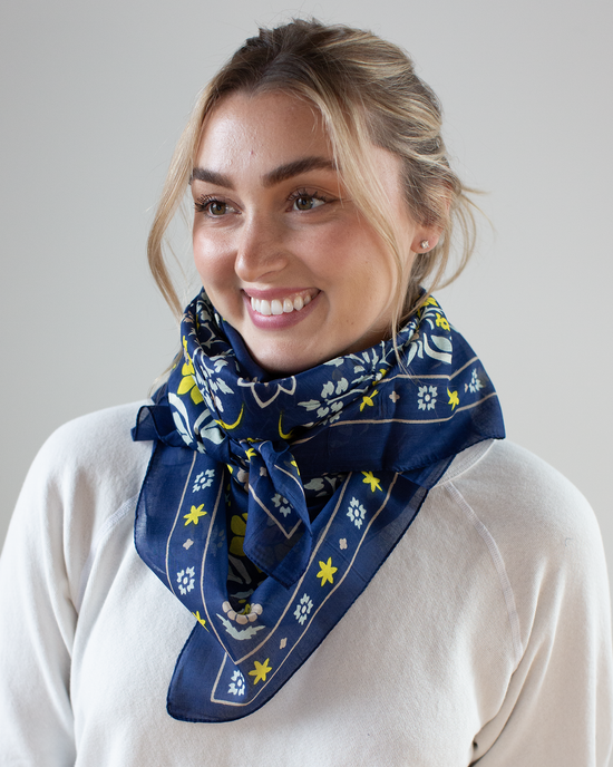 A person wearing a white sweater and a Maison Lecomte Flament Secret Garden Cotton Bandana in Navy smiles while looking to the side against a plain background.