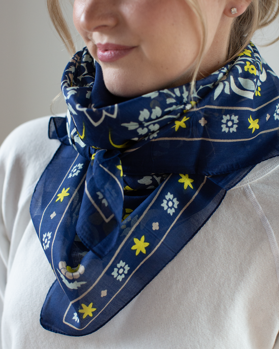 The woman is wearing a Maison Lecomte Flament Secret Garden Cotton Bandana in navy, showcasing yellow accents. Its delicately draped around her neck, complementing her white top as she smiles softly.