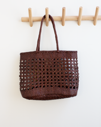The Eclipse Bag in Bruciato by Dragon Diffusion, featuring braided handles, hangs on a wooden peg rack against a white wall.