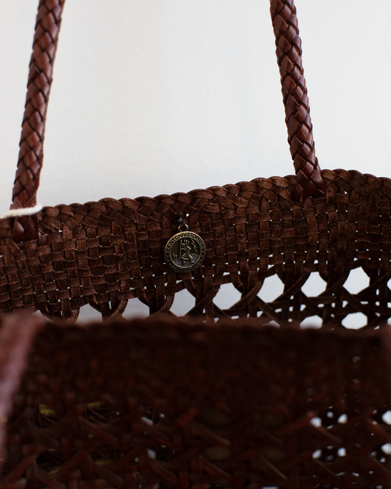 Close-up of the Eclipse Bag in Bruciato by Dragon Diffusion, a brown woven buffalo leather bag with braided handles and a round metallic emblem on the front.