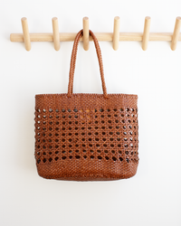 The Eclipse Bag in Tan by Dragon Diffusion, a brown woven tote with two handles, hangs on a wooden wall rack with multiple pegs.