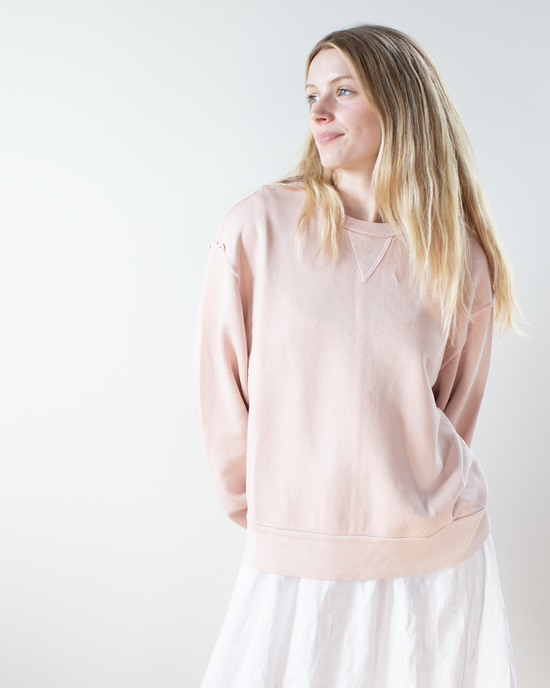 A woman with long blonde hair models the NSF Hayes Oversize Crew Sweatshirt in Rosy with a white skirt, standing against a plain white background and looking to the side.
