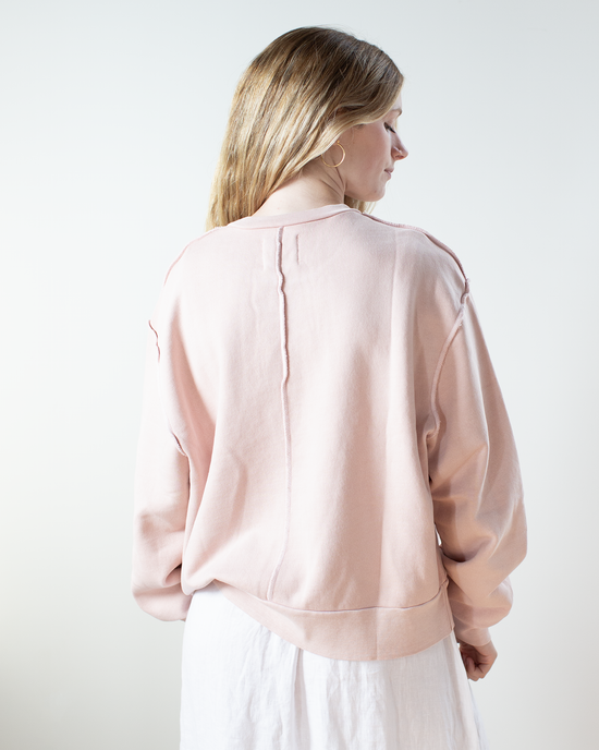 A woman with long light brown hair stands facing away, wearing the NSF Hayes Oversize Crew Sweatshirt in Rosy and a white skirt against a plain light background.