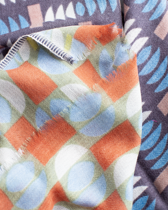 Close-up of two overlapping patterned fabrics: Épices Pop Geo Wool Silk & Cashmere Scarf in Mauve with blue, white, and beige geometric shapes, and another with orange, green, and blue circles and checks—both display rich texture.