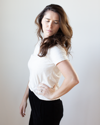 A woman with long brown hair stands sideways, looking down thoughtfully. She wears the Eli V Neck Top in Bisque by Velvet by Graham & Spencer and black pants, one hand on her hip against a plain light background.