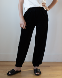 A person stands on a wood floor against a plain white wall, wearing Velvet by Graham & Spencer’s Essie Pant in Black, an elastic waistband jogger, paired with a white short-sleeve shirt and black slide sandals. One hand rests on their hip.