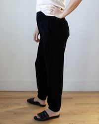 A person wearing a white shirt and the Velvet by Graham & Spencer Essie Pant in Black stands on a wood floor against a plain white wall, with one hand resting on their hip.