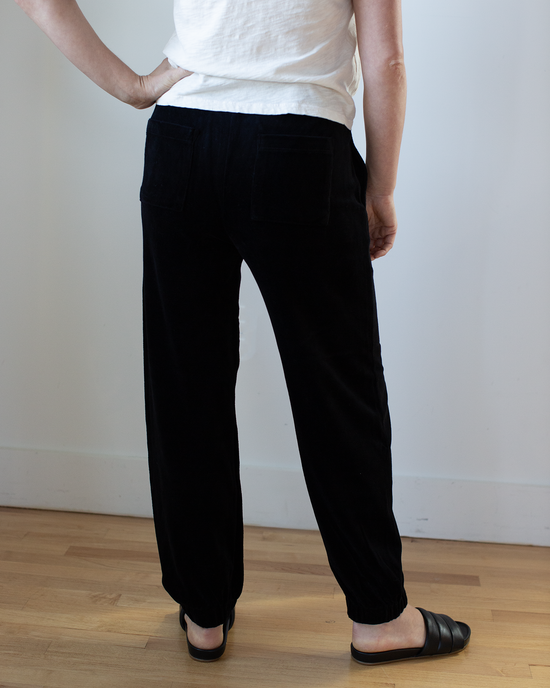 A person stands indoors with their back to the camera, wearing a white shirt and Velvet by Graham & Spencer’s Essie Pant in Black, paired with black slide sandals on a light wooden floor.