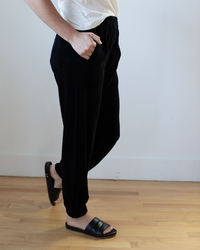 Model wears the Essie Pant in Black by Velvet by Graham & Spencer, featuring an elastic waistband and jogger style, paired with a white shirt and black slide sandals. Photo is cropped above the shoulders; shot indoors on a light wood floor.