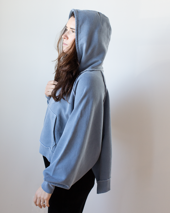 A person stands sideways against a plain light background, wearing the NSF Luma Oversize FT Poncho in Faded Indigo, gazing ahead with one hand holding the hoodie near their chest.