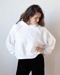 A woman with long brown hair wears the NSF Luma Oversize FT Poncho in Chalk and black pants, standing against a plain light background, looking down with one hand on her hip.