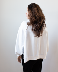 A woman with long, wavy brown hair faces away from the camera, wearing the NSF Luma Oversize FT Poncho in Chalk and black pants against a plain light background.
