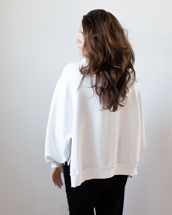 A woman with long, wavy brown hair faces away from the camera, wearing the NSF Luma Oversize FT Poncho in Chalk and black pants against a plain light background.