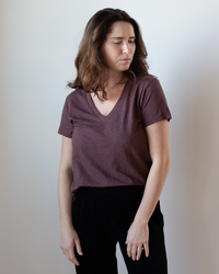 A woman with wavy brown hair wears the Velvet by Graham & Spencer Eli V Neck Top in Henna and black pants, standing against a plain light background and looking down with a neutral expression.