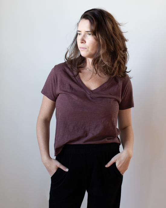 A woman with wavy brown hair wears the Eli V Neck Top in Henna by Velvet by Graham & Spencer and black pants, standing against a plain light background, looking left with her hands in her pockets.