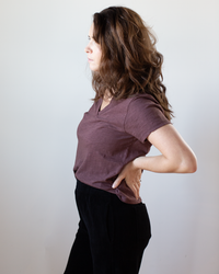 A woman with wavy brown hair stands in profile against a plain background, wearing the Velvet by Graham & Spencer Eli V Neck Top in Henna and black pants, one hand resting on her lower back.