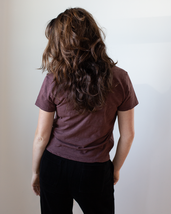 A person with long wavy brown hair faces away from the camera, wearing a Velvet by Graham & Spencer Eli V Neck Top in Henna and black pants against a plain light-colored background.