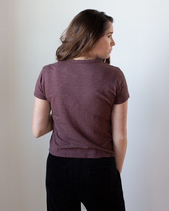 A woman with long brown hair stands facing away, wearing the Velvet by Graham & Spencer Marie S/S Crop Top in Henna and black pants, her right hand in her pocket against a plain white background.