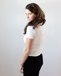 A woman with long brown hair, wearing the Enati S/S Crew Neck Top in Ecru by Velvet by Graham & Spencer and black pants, stands turned slightly away from the camera, looking down neutrally against a plain light background.