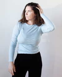 A woman with long brown hair wears the Velvet by Graham & Spencer Mauri L/S Crew Neck Top in Ceramic, paired with black pants, looking left and touching her head against a plain light background.