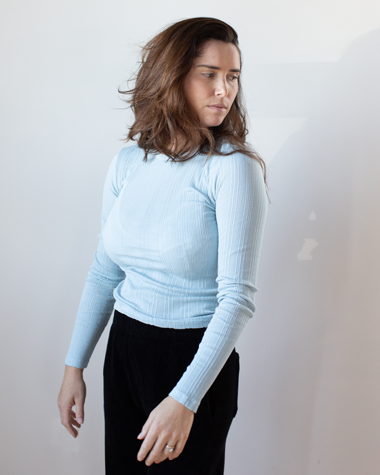 A woman with long brown hair wears the Mauri L/S Crew Neck Top in Ceramic by Velvet by Graham & Spencer and black pants, standing against a white background and looking to her left with a neutral expression.